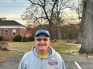 Tony Fiore's smiles while wearing a blue Trees Atlanta Certified Volunteer ball cap. Behind him are trees with no leaves and sunset. 