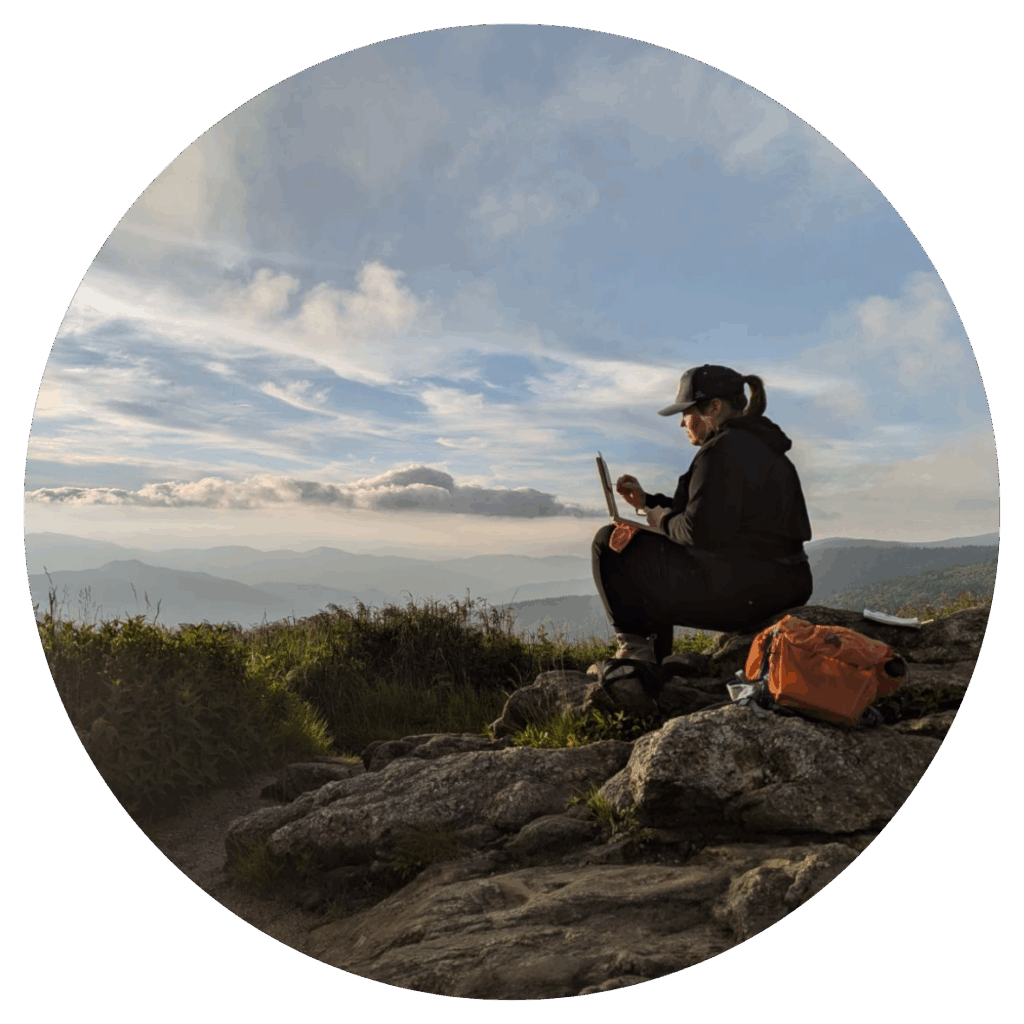 Rounded image shows a woman sitting on rocks on top of a mountain looking out at a cloudy sky with a mountain range in the background. 