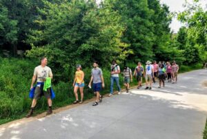 A group of people walk in a line along a path with trees behind them. The person at the front looks back to see speak to the group, he is giving a tour. 