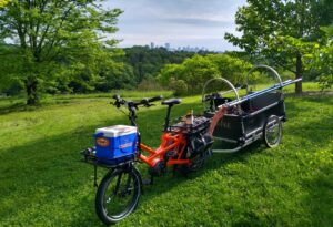 A bicycle stands with a heavy-duty trailer attached to the back with tools inside. In the front, a basket holds a cooler. The bike its in a grassy lawn. Behind the bike are trees and a distant skyline.