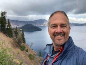 A man smiles in the foreground. Behind him is a vast lake and cliffside with sparse trees. 