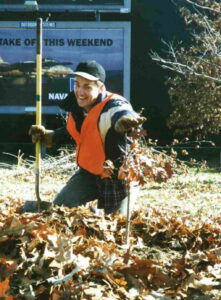 A man kneels on the ground in a pile of fallen leaves. He holds a shovel and a recently planted small tree is in front of him. 