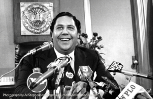A man stands smiling behind a podium with various microphones pointed towards him. In the background you see the seal for the City of Atlanta.